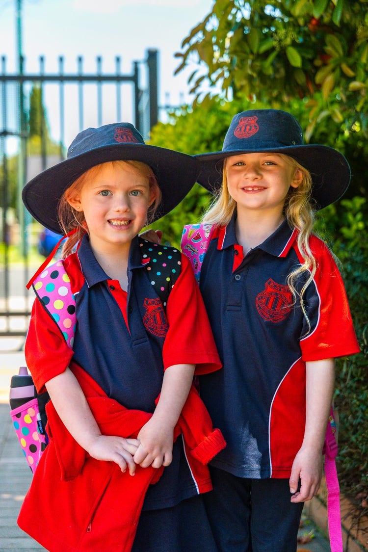 Two girls standing near the front gate with their hats on and backpacks on their backs ready for a great day.