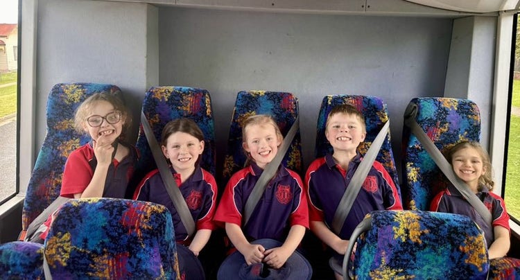 Five students sitting in the back row of a bus with their seat-belts on.