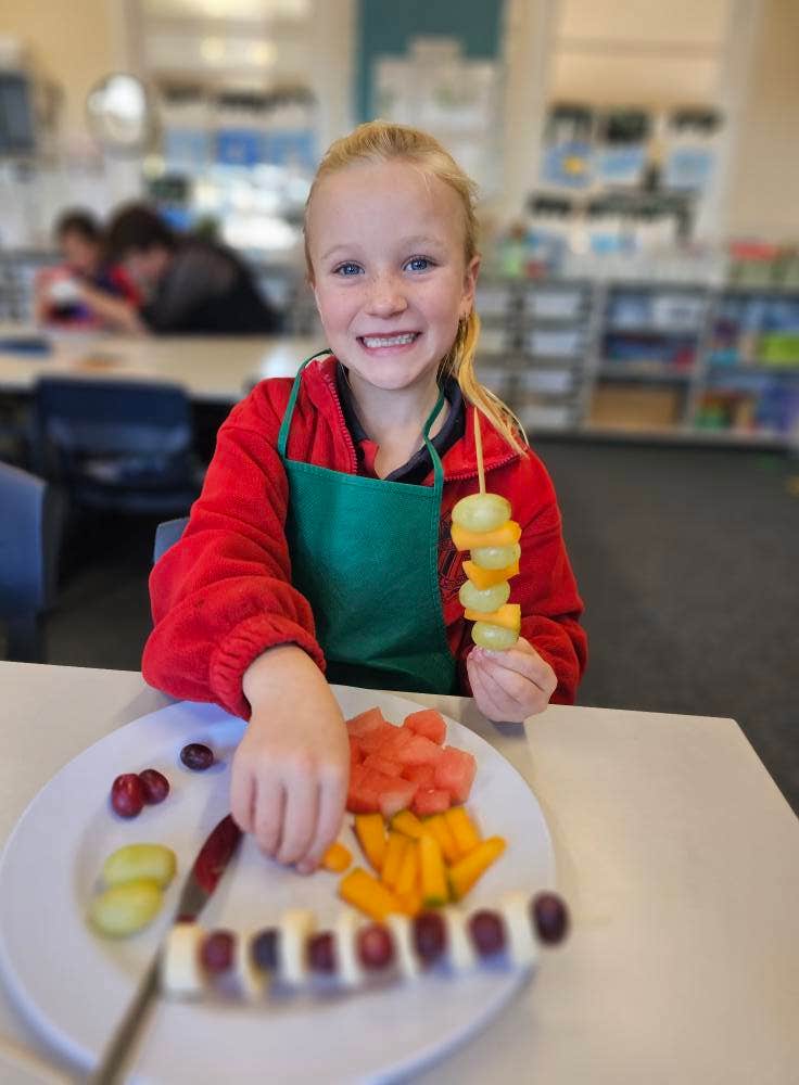A student making fruit kebabs with a green apron on.